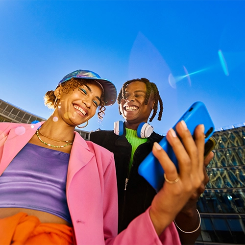 Mann und Frau in Freizeitkleidung halten Smartphones und lächeln; urbanes Umfeld bei blauem Himmel.