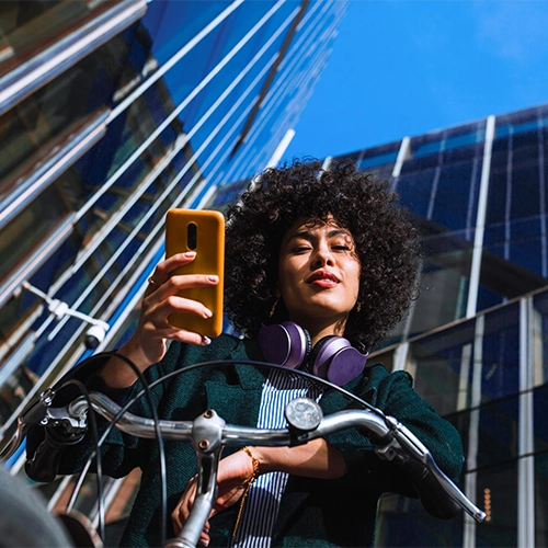 Frau sitzt auf einem Fahrrad in Freizeitkleidung, hält ein oranges Smartphone; Gebäude und blauer Himmel im Hintergrund.