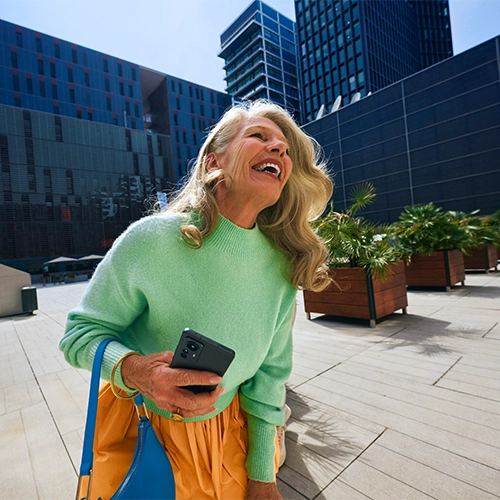 Smiling woman in a green sweater with orange pants and blue bag, holding a cell phone surrounded by an urban background.