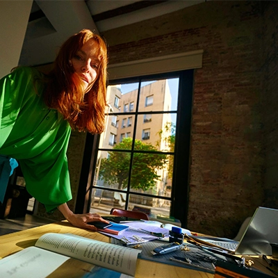 Woman in a green shirt standing at a desk with a laptop and office supplies; building and tree visible through the window.