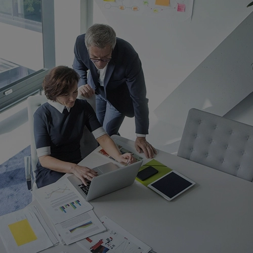 Man and woman in business attire talk in an office in front of a laptop; desk with documents and pens.
