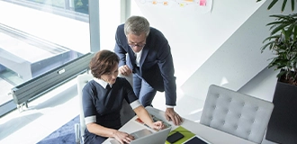 Man and woman in business attire in an office talking in front of a laptop.
