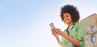 Woman in casual clothes smiling at her cell phone; part of a building and blue sky in the background.