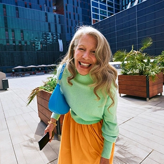 Woman in casual clothes smiling with smartphone in front of urban background with plants.