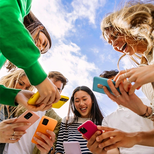 Six smiling people with colorful smartphones in front of a blue sky with clouds.